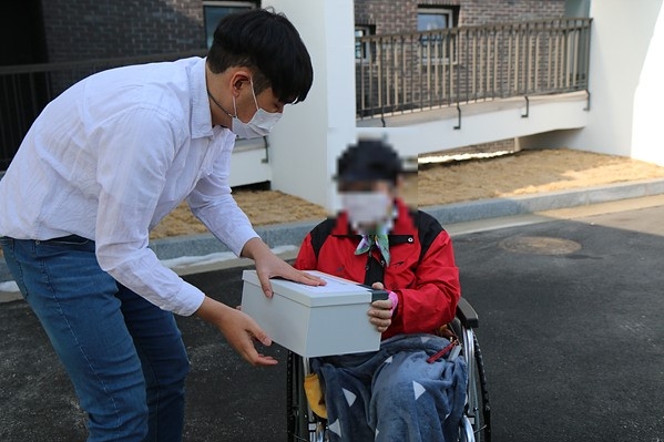 이천시장애인복지관 근무가족이 휠체어 장애가족에게 설날을 맞이하여 무언가가 들어있는 상자를 전달하고 있다/ 이용인의 얼굴에는 모자이크 처리가 되어 있다