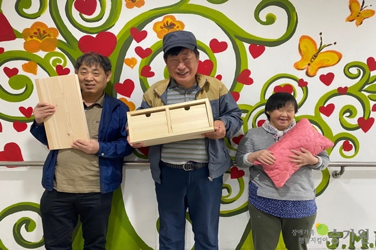 각자가 만든 편백나무 작품을 들고 사진을 찍고 있는 세명의 장애가족 - 장애가족 행복지킴이 승가원 ci 삽입