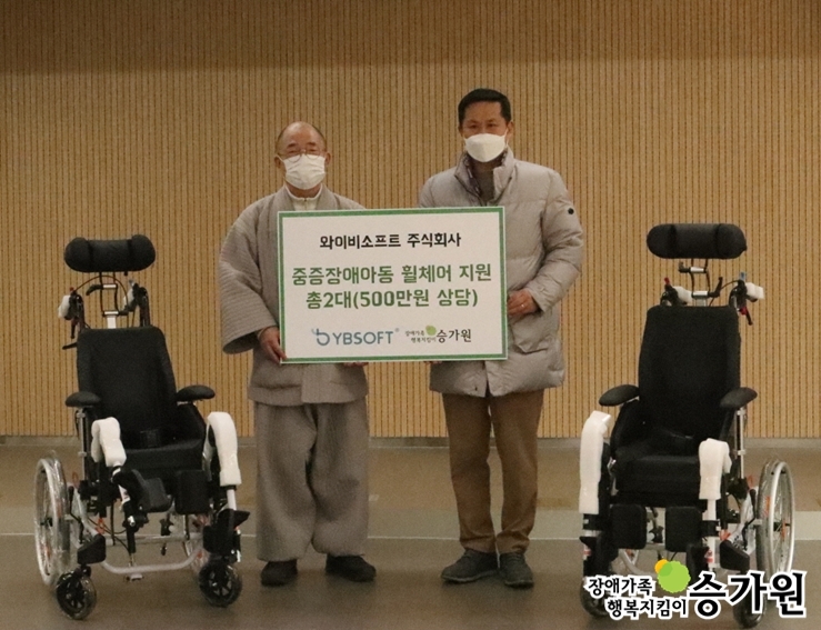 국장스님과 주식회사와이비소프트 대표이사가 휠체어 전달식을 하고 있는 모습. 양옆에 휠체어가 한 대씩 놓여있음.