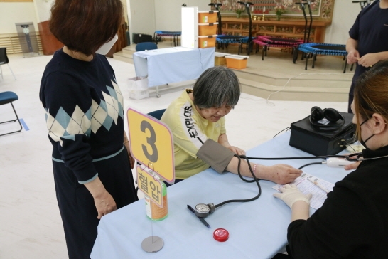 테이블 위에 3/혈압이라고 적혀져 있다. 병원직원이 여성 장애가족의 혈압을 재고 있는 모습이고 직원이 뒤에서 지켜보고 있다