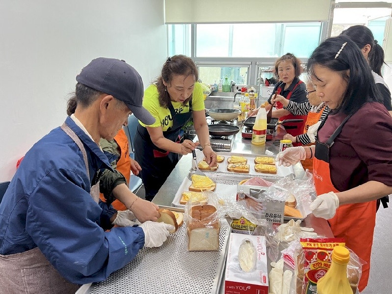 참여자들이 성북장애인복지관에 모여서 요리활동을 진행하고 있다. 주민들은 샌드위치를 만들기 위해 장갑을 끼고 있거나 뒤집개를 들고 있으며 모두가 앞치마를 매고 있다