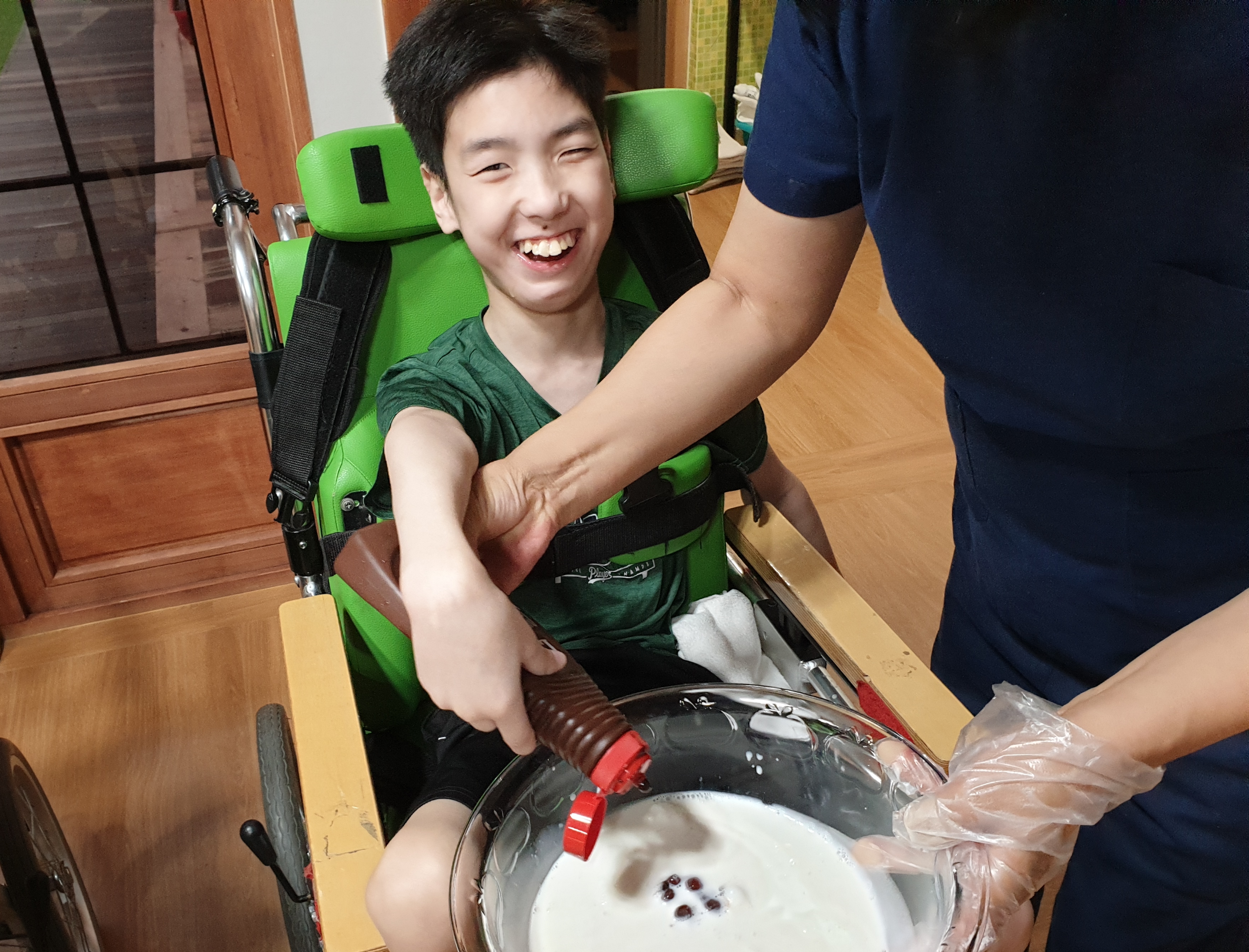 휠체어를 타고 있는 남자 장애아동이 근무가족과 함께 빙수만들기를 하고 있는 사진