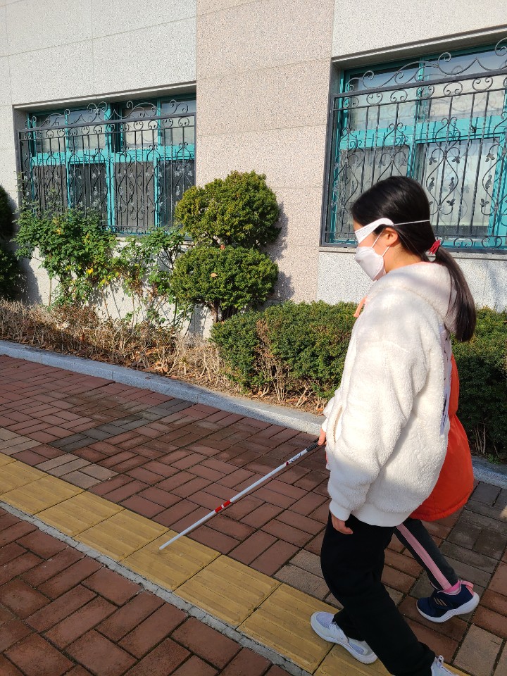 시각장애체험 안경을 쓰고 흰지팡이를 사용해보고 있는 학생 1명, 친구에게 방향을 안내해주는 학생 1명이 학교 건물 앞을 함께 걸어보고 있다 