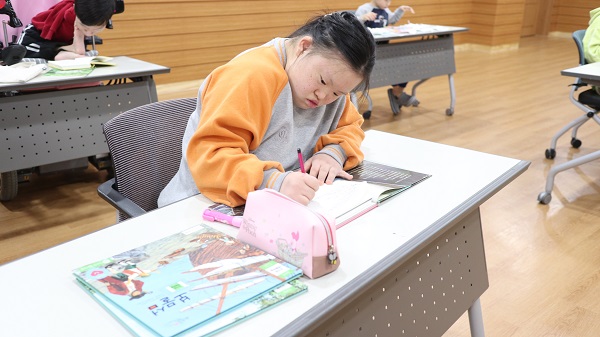 여자 장애아동이 한 손으론 책을 누르고 한 손으론 연필을 들고 글씨를 쓰고 있다