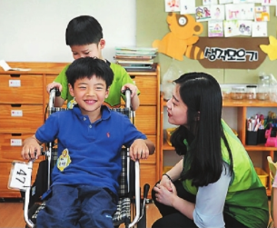 초등학생 남자아이가 휘체어를 타고 뒤에서 친구가 밀어주며 장애체험을 하고 있고 옆에는 사회복지사가 앉아있는 모습