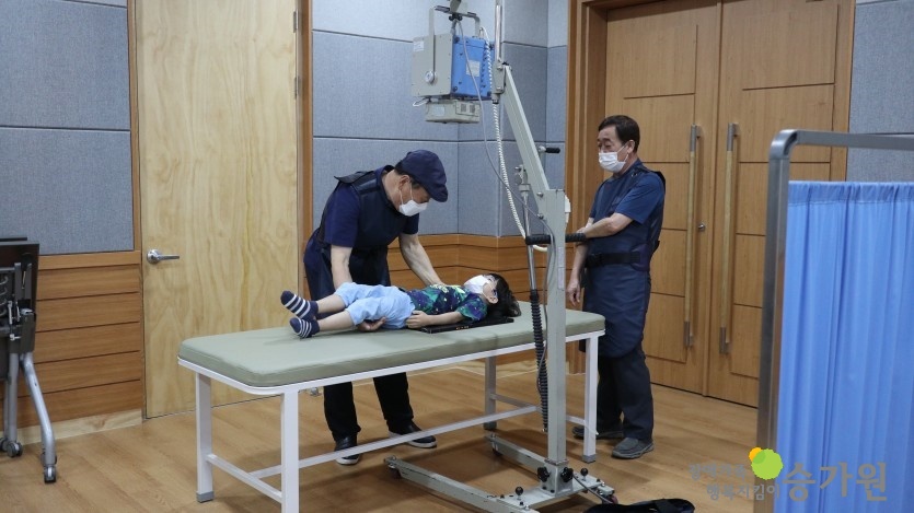 엑스레이를 찍기 위해 기계 위에 누워있는 남자 아동과 이를 도와주고 있는 두 명의 남자 선생님들의 모습/오른쪽 하단에 장애가족행복지킴이 승가원 CI로고 삽입