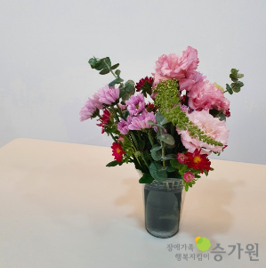 책상 위에 화분 한 개가 놓여져 있다. 보라빛 꽃과 분홍색 꽃, 빨간색 꽃이 조화롭게 꽂아져 있는 화분의 모습이다./오른쪽 하단에 장애가족행복지킴이 승가원 CI로고 삽입