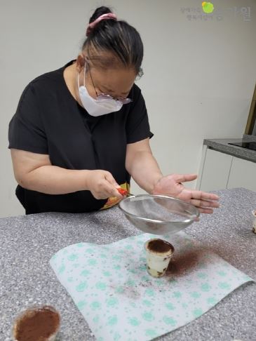 여성 장애가족이 지인에게 선물할 티라미수 컵케이크를 만들기 위해 케이크에 초코파우더를 뿌리고 있는 모습/오른쪽 상단에 장애가족행복지킴이 승가원 CI로고 삽입
