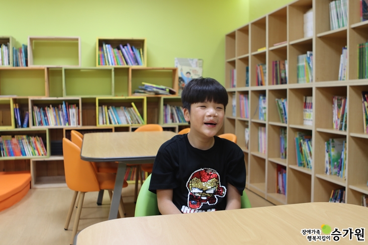 박철우 아동이 초록색 의자에 앉아 해맑게 웃고 있는 모습/오른쪽 하단에 장애가족행복지킴이 승가원 CI로고 삽입