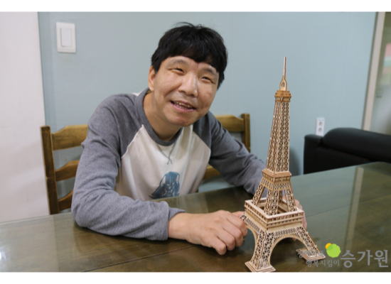 남자 장애가족이 직접 만든 에펠탑 조립 키트를 테이블에 올려놓고 카메라를 보며 웃고있다./오른쪽 하단에 장애가족행복지킴이 승가원 CI로고 삽입