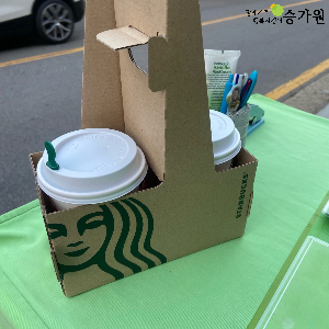 초록색 책상 위에는 스타벅스 종이캐리어에 담긴 따뜻한 음료 두잔이 놓여있음. 오른쪽 상단 위에 장애가족지킴이 승가원 ci삽입