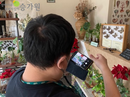 나비표본과 각종 식물과 꽃이 있으며 이를 남성 장애가족이 휴대전화 카메라를 이용해 사진을 찍고 있다. 장애가족행복지킴이CI.