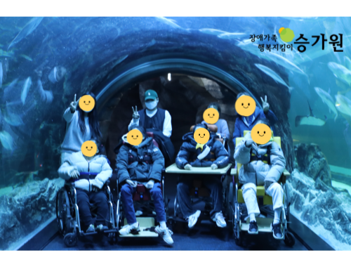 상단 오른쪽에는 장애가족 행복지킴이 승가원ci/ 물고기가 보이는 투명한 둥근 유리관 통로에 담당교사와 휠체어에 앉은 장애가족 총 8명이 찍힌 사진