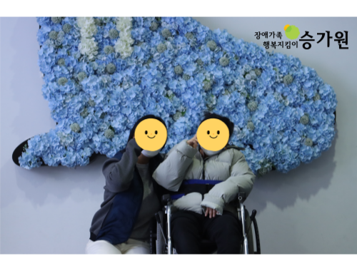  상단 오른쪽에는 장애가족 행복지킴이 승가원ci/ 하늘색 꽃으로 만든 물개를 배경으로 사진을 찍고 있는 담당교사와 장애가족의 모습
