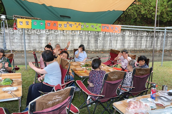 연꽃문수캠프 맛있게~ 신나게~ 행복하자 라고 적힌 종이현수막 앞에 복지타운 이용인들이 캠핑용품을 펼쳐놓고 바비큐 파티를 즐기고 있다.