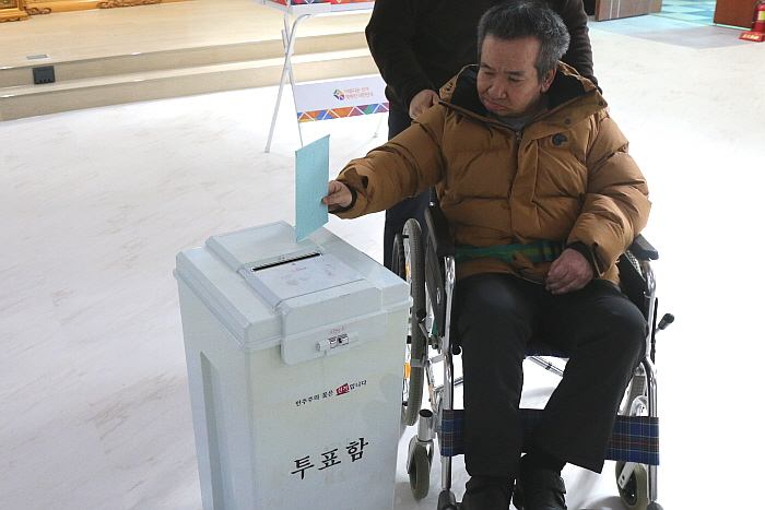 투표함에 투표용지를 넣는 휠체어에 앉은 남성 장애가족, 고동색 패딩을 입고 있다.