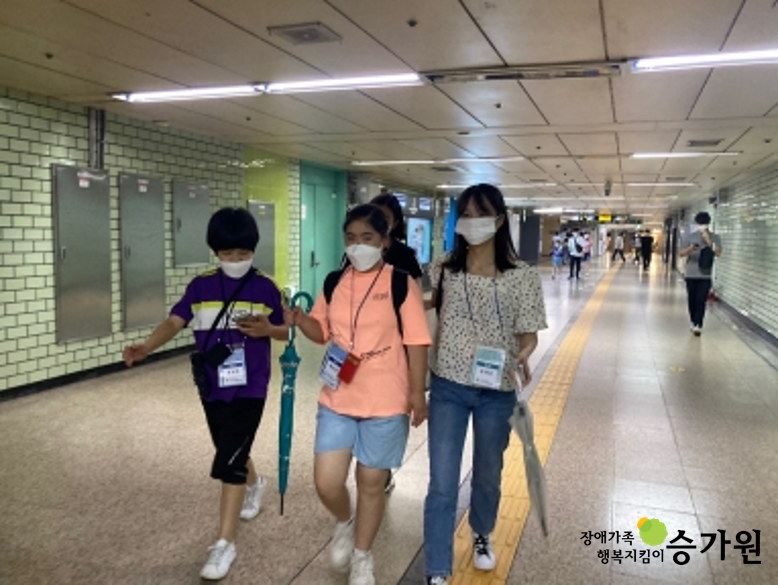 마스크를 착용한 3명의 장애 청소년이 나란히 서서 지하철역 통로를 걷는 모습. 왼쪽부터 차례로 네이비색 상의, 형광 주황색 상의, 검정색 무늬가 있는 흰색 상의를 입고 있으며, 오른쪽의 두 명은 손에 우산을 들고 있다. 또한, 모두 참여자 명단 목걸이를 메고 있다. 우측 하단 장애가족행복지킴이 승가원 CI 삽입