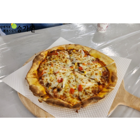 직업 적응 훈련생들이 직접 만든 피자 1개가 책상 앞에 놓여져 있습니다. 피자는 치즈가 많아보이며, 빨간색 야채도 들어 있습니다. 좌측 상단 ci 승가원 삽입