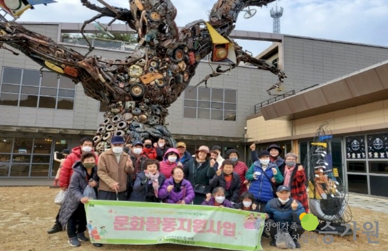 약 17명의 장애가족들이 문화활동지원사업 현수막을 들고 단체사진을 찍고 있는 모습, 뒤에는 꾸며진 나무가 있다. 장애가족행복지킴이 승가원 ci  로고 삽입!