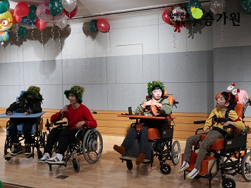 휠체어를 탄 장애아동 4명이 크리스마스 트리모양의 모자를 쓰고 크리스마스를 즐기고 있는 사진. 천장에는 알록달록한 크리스마스 풍선이 매달려 있다. 우측 상단에 장애가족행복지킴이승가원ci 삽입.