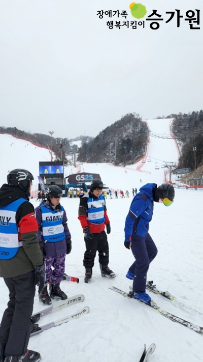 새하얀 눈으로 뒤덮인 스키장에 세 명의 남성이 FAMILY라고 적힌 흰색, 파란색 줄무늬 문양의 조끼와 스키복을 착용하고 있다. 왼쪽에 서 있는 세 명의 남성은 오른쪽에 파란색 외투를 입고 형광색 고글을 착용한 남성의 스키 장비를 다루는 모습을 바라보고 있다. 오른쪽 상단 장애가족 행복지킴이 승가원CI
