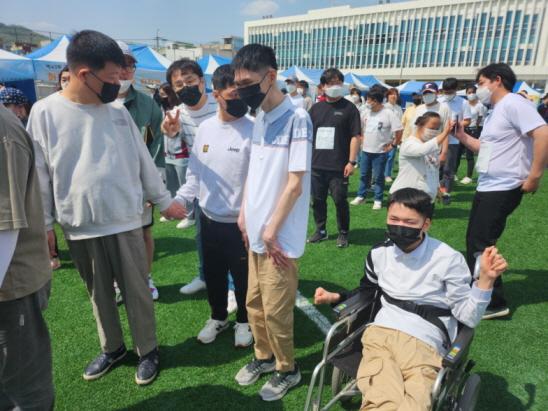 야외의 잔디로 된 운동장에서 휠체어를 탄 장애가족, 서로 손을 잡고 있는 장애가족들과 복지사들이 줄을 맞춰 길게 서 있는 모습.
