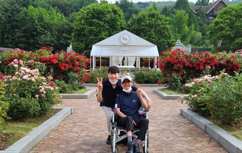 휠체어를 탄 장애가족 1명, 장애가족과 함께 사진을 찍은 근무가족 1명의 모습. 뒷편에는 장미꽃들이 가득하다. 두 사람은 모두 한 손으로 브이자를 만들어 보이며 활짝 웃고 있다.