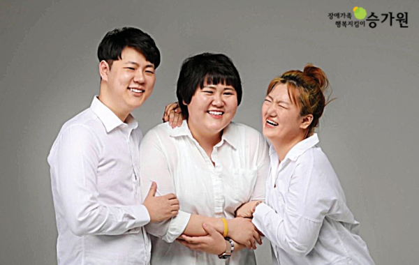 오른쪽 상단에 장애가족행복지킴이 승가원ci삽입. 하얀셔츠와 청바지를 맞춰입은 김다영 후원가족님 삼남매가 서로를 껴안으며 환하게 웃고 있음. 두 동생들 사이에 김다영 후원가족님.