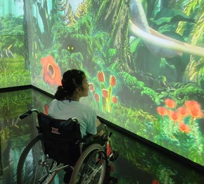 장애아동이 휠체어에 앉아 스크린을 바라보고 있다. 스크린에는 정글 같은 풀숲과 튤립 등의 꽃이 보인다.