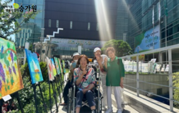 왼쪽 상단에 장애가족 행복지킴이 승가원CI 삽입 / 그림들이 왼쪽 화단에 세워져있고, 휠체어를 탄 장애가족과 여성 3명이 환히 웃으며 손 하트를 하고 있음.