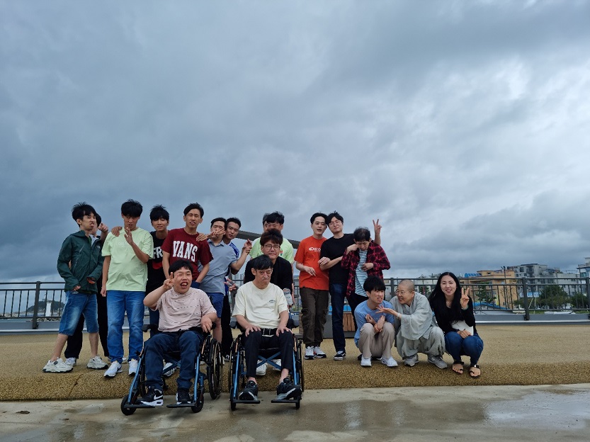 16명의 장애가족들과 직원 1명, 스님 한 분이 해변공원에서 단체사진을 찍은 모습. 앞줄은 휠체어를 타고 있고, 뒷줄은 한줄로 서서 프이포즈를 하고 있다.