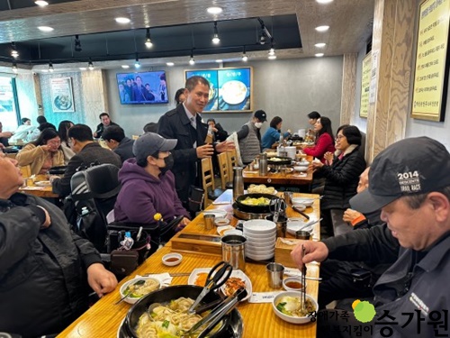 한 남성이 검은 외투를 입은 채 식당에 서서 테이블에 앉아 있는 사람들에게 무언가를 이야기하고 있다. 테이블 위에는 김치와 만두전골, 앞 접시 등의 음식들이 있다. 오른쪽 하단 장애가족 행복지킴이 승가원ci