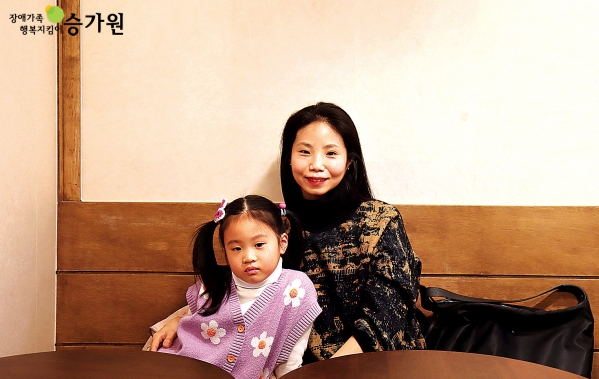 연보라색 조끼를 입은 삐삐머리 여자아이의 옆에 앉아 미소를 짓고 있는 긴 생머리의 여성/왼쪽 상단 장애가족행복지킴이 승가원ci