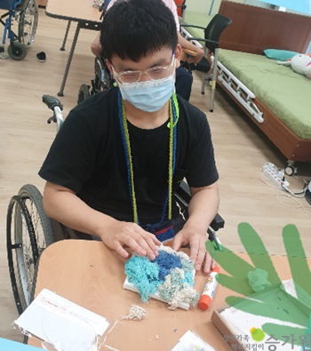 휠체어에 앉아 푸른색 스칸디아모스 액자를 만들고 완성한 남자 장애가족의 모습/오른쪽 하단에 장애가족행복지킴이 승가원 CI로고 삽입