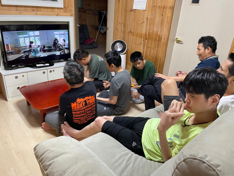 7명의 남성 장애가족들이 생활실에 앉아서 TV에서 나오는 인권교육 영상을 시청하고 있다. 장애가족행복지킴이 승가원CI