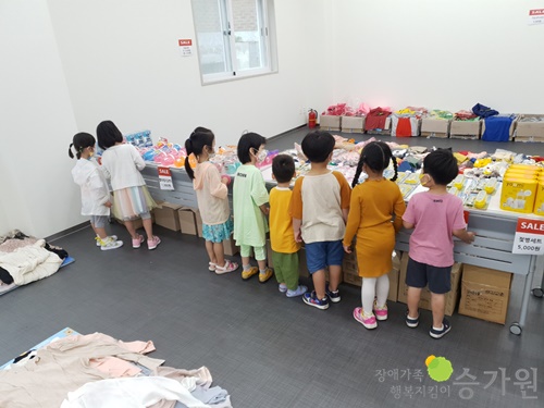 8명의 어린아이들이 유아용품 앞에서 물건을 구경하고 있다./오른쪽 하단에 장애가족행복지킴이 승가원 CI로고 삽입