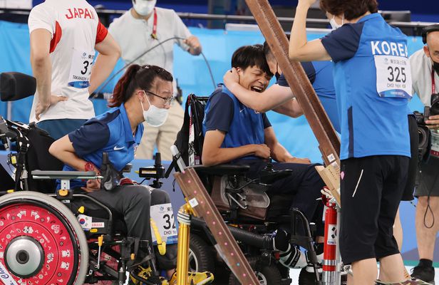 휠체어에 앉은 남성 패럴림픽 선수가 기쁜 표정으로 남성 코치와 끌어안고 있다. 그 오른쪽에는 여성 코치가 뒷 모습을 보이며 서있다. 왼쪽에는 어성 패럴림픽 선수가 휠체어에 앉아있는 모습이다. 