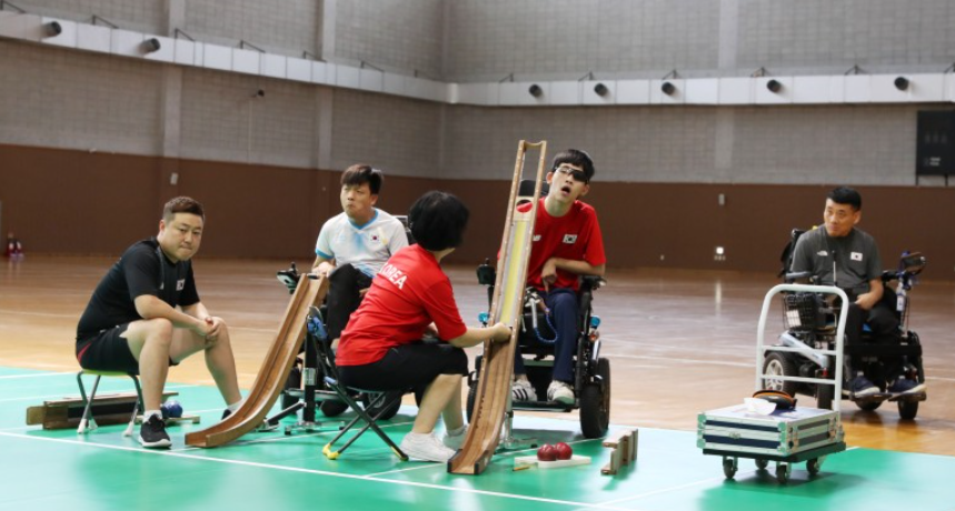 패럴림픽 보치아 선수들과 코치들의 모습. 3명의 남성 패럴림픽 보치아 선수들이 휠체어에 앉아있다. 그 앞으로 남성 코치와 여성 코치가 의자에 앉아있는 모습이다.