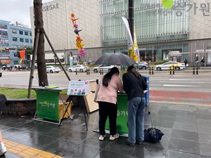 천안터미널에서 캠페인을 진행하는 와중에 갑자기 비가 와서 여성 사회복지사가 우산을 들고 시민 분에게 행복나눔캠페인을 설명하고 있는 모습. 우측상단에 장애가족행복지킴이CI.