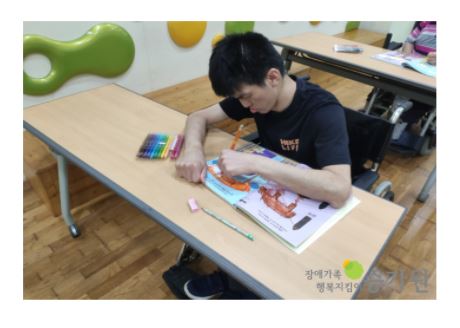 한 남성 장애가족이 책상에 앉아서 주황색 색연필을 이용해 색칠연습을 하고 있다. / 장애가족 행복지킴이 승가원 ci 삽입