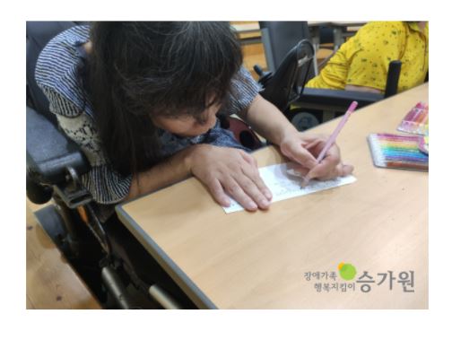 여성 장애가족이 책상에 앉아 분홍색 연필을 이용하여 편지를 쓰고 있다. / 장애가족 행복지킴이 승가원 ci 삽입