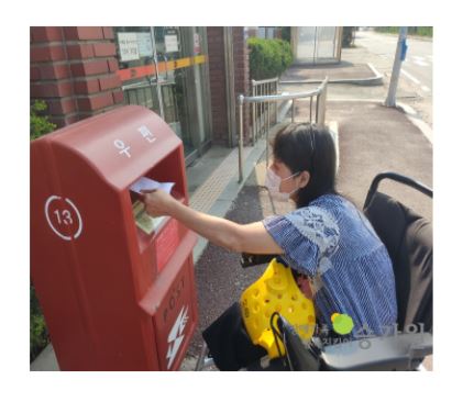 휠체어에 앉아 빨간 우체통에 편지를 넣고 있는 여성 장애가족의 모습/장애가족 행복지킴이 승가원 ci 삽입