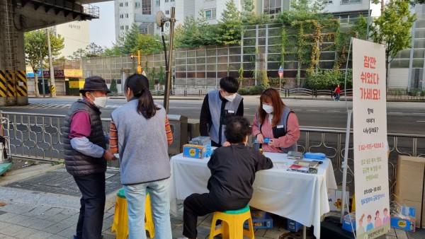 세 명의 사회복지사들이 두 명의 성북구 주민들을 만나 거리에서 캠페인을 진행하고 있다. 캠페인 현장에 게시판과 테이블이 준비되어 있다.