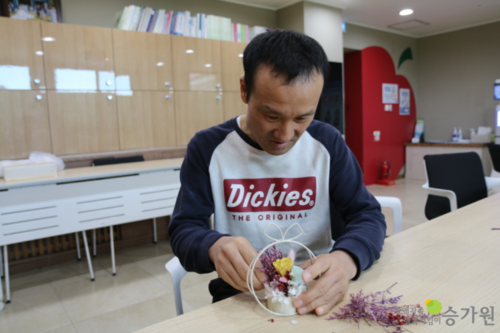 승가원자비복지타운 자비마을 이용인 한 명이 비대면키트 만들기로 꽃을 이용한 소품을 만드는 모습. 우측 하단에 장애가족행복지킴이 승가원 ci 삽입.
