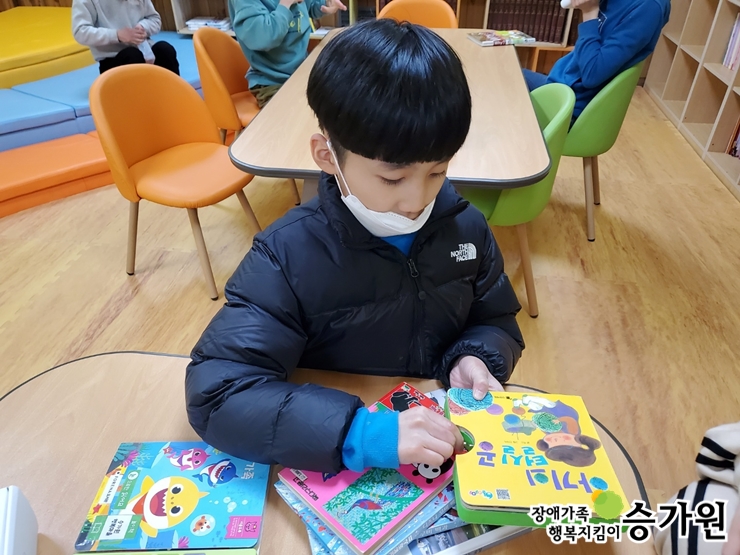 김동재 장애아동이 테이블 위에 7개의 책을 쌓아 놓았다. 그 중 아기와 털실공이라는 책을 읽기위해 손을 올려두었다. 왼쪽 아래 장애가족 행복지킴이 승가원CI 삽입
