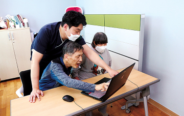 장애가족 두 명이 책상에 앉아 노트북을 보고 있다. 그 중 한명과 뒤에 서 있는 근무가족이 같이 노트북을 향해 손가락으로 가리키고 있다. 