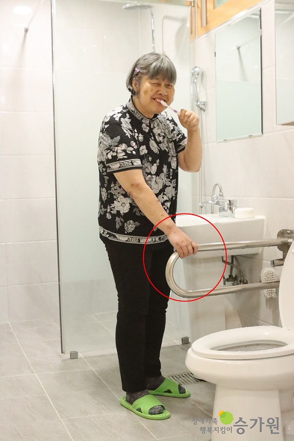 여성장애인이 화장실 세면대 앞에서 한손으로는 안전손잡이를 잡고 한손으로는 칫솔을 들고 양치를 하고 있다.