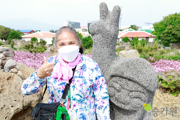 활짝핀 분홍꽃과 브이하는 석상을 배경으로 손가락 하트를 하며 웃고있는 여자 장애가족의 모습/ 장애가족행복지킴이 승가원ci