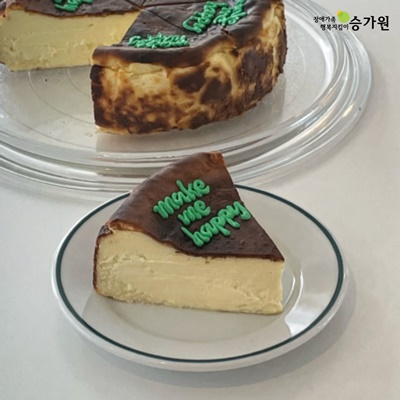 치즈 홀케익이 놓여져있고 한 조각은 따로 앞 접시에 놓여져있음. 케이크 위에는 초록색으로 'make me happy'라고 적혀져있음. 오른쪽 위 장애가족 행복지킴이 승가원CI