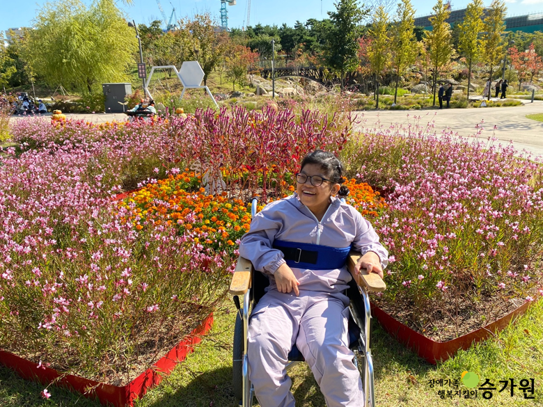 휠체어를 탄 장애아동이 미소를 짓고 있는 모습. 가을에 핀 꽃들을 배경으로 하여 사진을 찍고 있다. 오른쪽 하단 장애가족 행복지킴이 승가원 CI